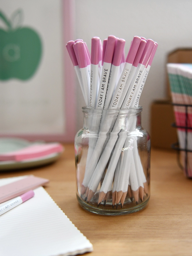 A glass jar full of white pencils with pink tips stands on a wooden desk surrounded by various stationery items.