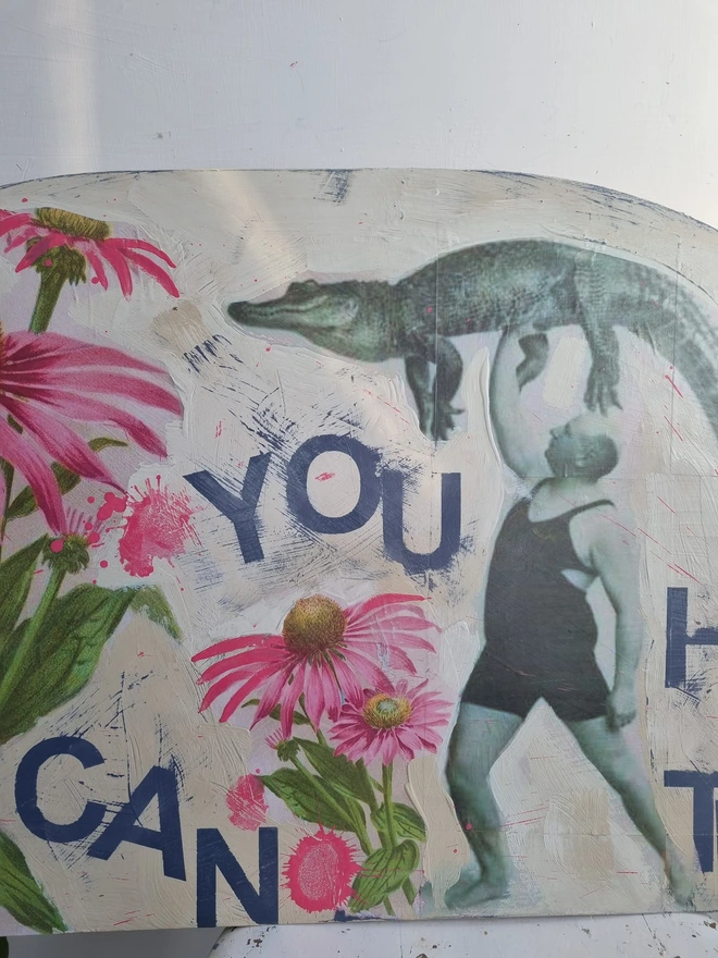 Art panel in a crescent shape made from a piece of vintage table top. Decorated with decoupage images of a man holding a crocodile aloft and pink flowers. The text reads 'you can do hard things'. The colours are cream with splashes of pink