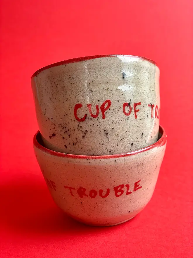 'Cup Of Trouble' Mug, two ceramic mugs that say 'Cup Of Trouble' on them stacked together in front of a red background. 