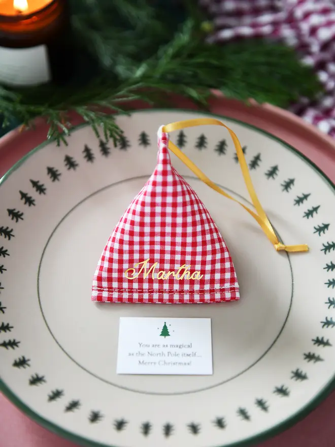 A red gingham fabric fortune cookie with gold personalised name “Martha” sits on a Christmas dinner plate, ready as a handmade festive place setting with a hidden Christmas wish card beneath.