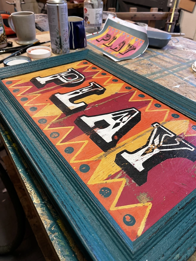 colourful play sign
made from reclaimed wood with a teal sign, hand painted fairground lettering and with orange pink and yellow stripes, with orange and yellow zig zag patterns  top and bottom