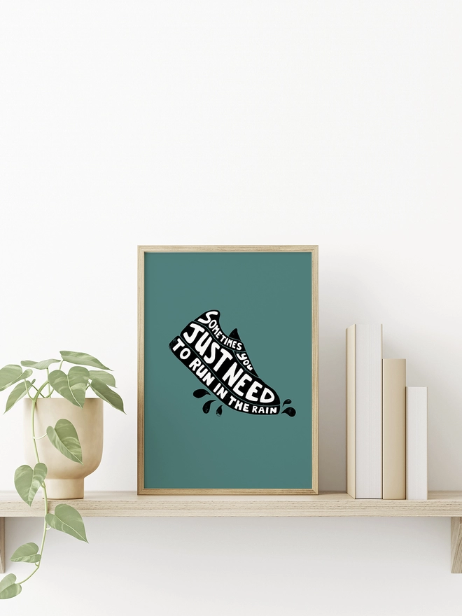 A bright print in a eooden frame sitting on a wooden shelf. The print is green and has typographic words making the outline of a running shoe. The words read: Sometimes You Just Need To Run In The Rain.