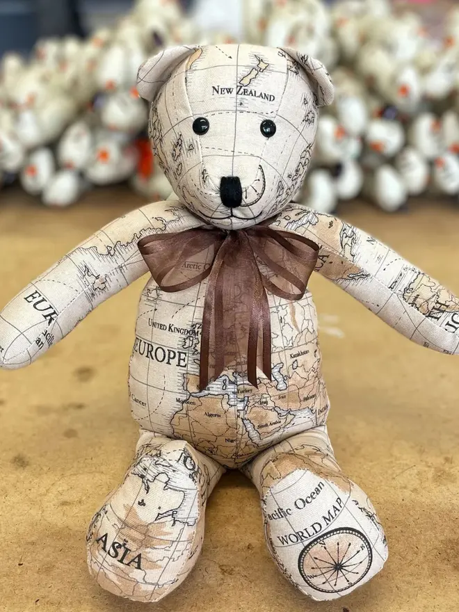 Teddy bear made from map-print fabric sitting upright on a wooden table, wearing a brown bow.