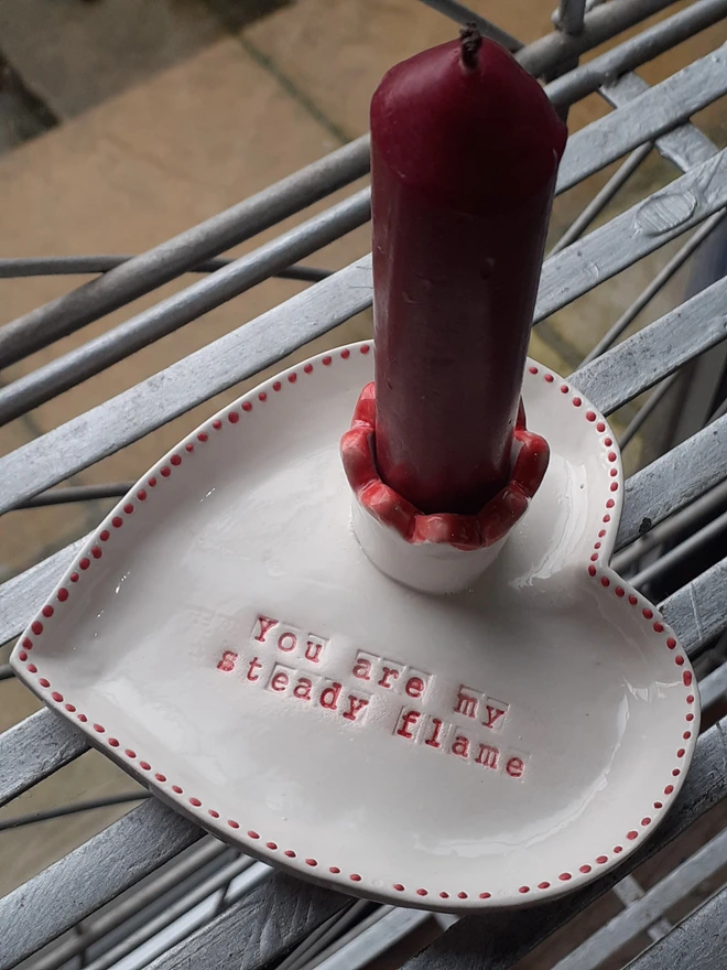 Glossy white heart-shaped ceramic taper holder with red decorative dots around the edge and a scalloped red candle cup, Valentine’s gift idea.