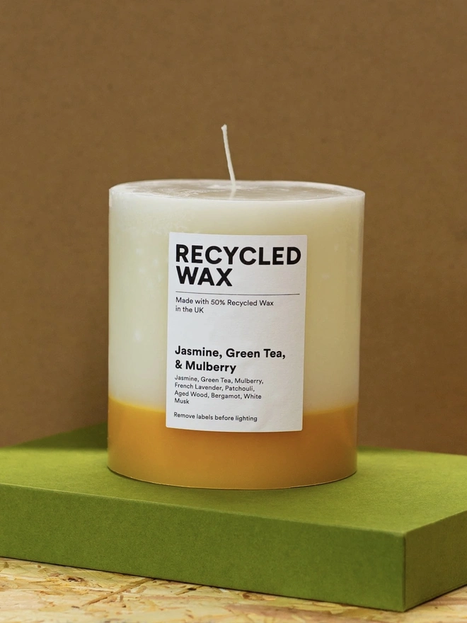 Jasmine, Green Tea & Mulberry Candle (Set Of 1 Or 3 Wicks). the jasmine, green tea and mulberry candle stands on a green box against a brown background, showing its single wick and cream wax with a golden yellow base. a white label reads "recycled wax, made with 50% recycled wax in the uk", followed by the scent name and note list of jasmine, green tea, mulberry, french lavender, patchouli, aged wood, bergamot and white musk. the candle's two-tone colouring and visible drip texture give it a rustic, hand-poured look.
