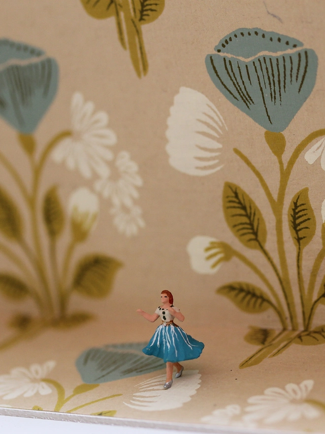 Miniature scene in an artbox showing a tiny lady in floral dress almost disappearing against matching floral wallpaper - close up