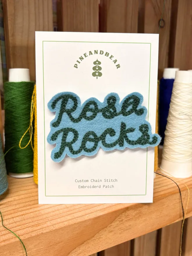 A chain stitched embroidered patch in teal and green spelling the word Rosa Rocks, on a white presentation card