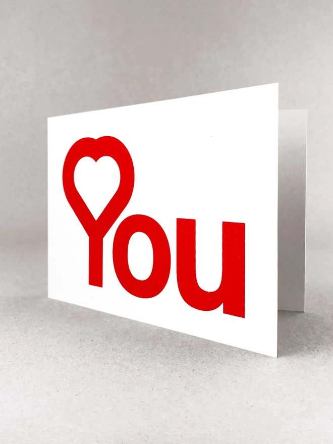 A heart in the Y of You, reads Heart You, screenprinted in red on a white landscape card. Stood on a white backdrop, casting a light grey shadow.