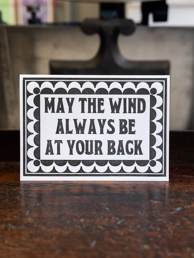 Letterpress card with text design "May The Wind Always Be At Your Back," surrounded by art deco style border of repeated semicircles 