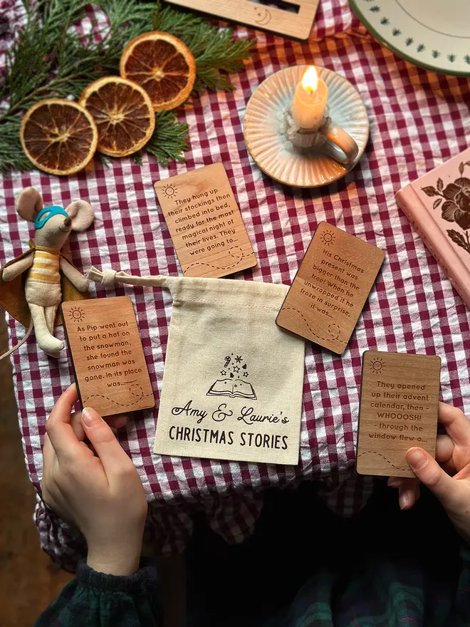 A personalised cotton bag labelled “Amy & Laurie’s Christmas Stories” on a red gingham table, surrounded by four engraved wooden story prompt cards, dried oranges, greenery, a candle, and a toy mouse.