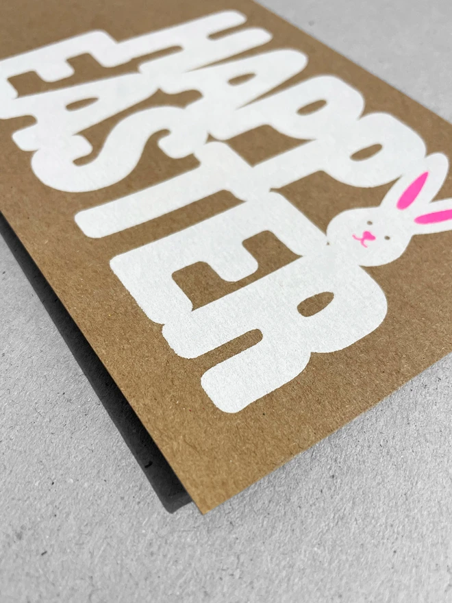 A close up of a kraft card on a grey background. The words Happy Easter are screen printed in white ink. The letter Y of happy forms the shape of a white bunny. Neon pink ink is printed over the top to create the ears and the classic twitchy bunny nose.