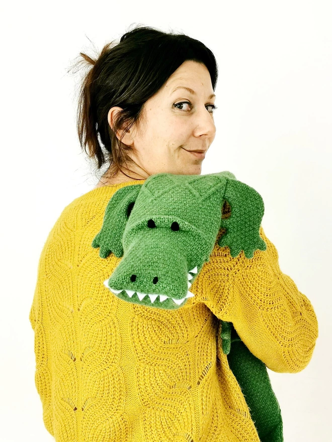 A woman with her back to us is holding a cuddly crocodile hot water bottle over her shoulder. The crocodile is looking cheekily at the camera and the woman is looking over her shoulder smiling.