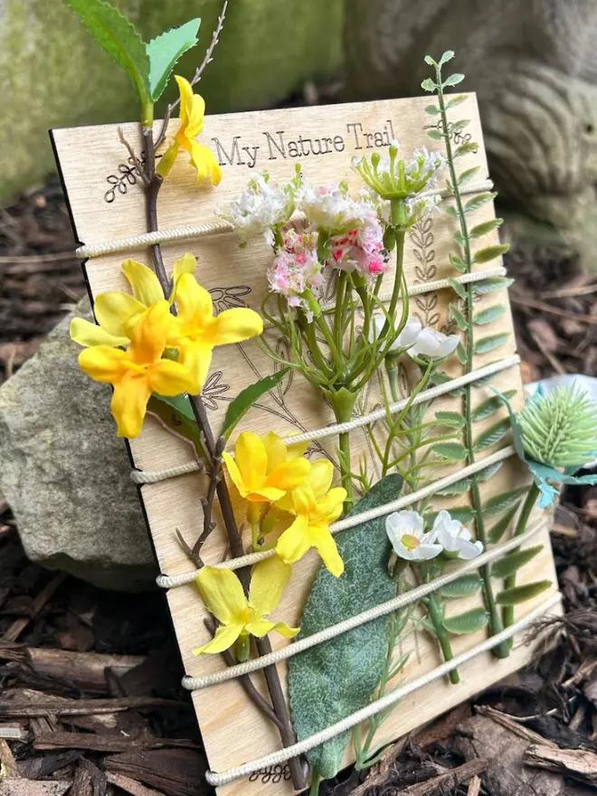Birch wood nature trail weaving board decorated with artificial wildflowers and leaves, held in place by cream-colored cord, displayed outdoors against mulch and stones.