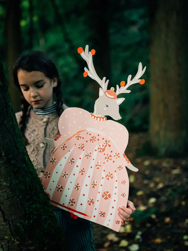 Extra Large Deer Deer Lady Wall Puppet. a girl in a woodland setting looks down at the extra large deer lady wall puppet, held up beside a mossy tree trunk. the deer wears a small orange cap between its painted antlers dotted with orange pompoms, above a full pink dress patterned with white polka dots, striped panels and peppermint-style motifs in orange and cream. the dark, forested backdrop and soft, muted lighting give the puppet a whimsical, storybook charm.
