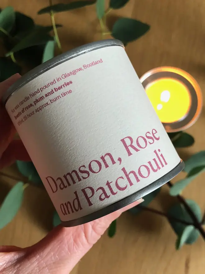 Damson, Rose & Patchouli Candle, held in a hand, displayed against a wooden background. 