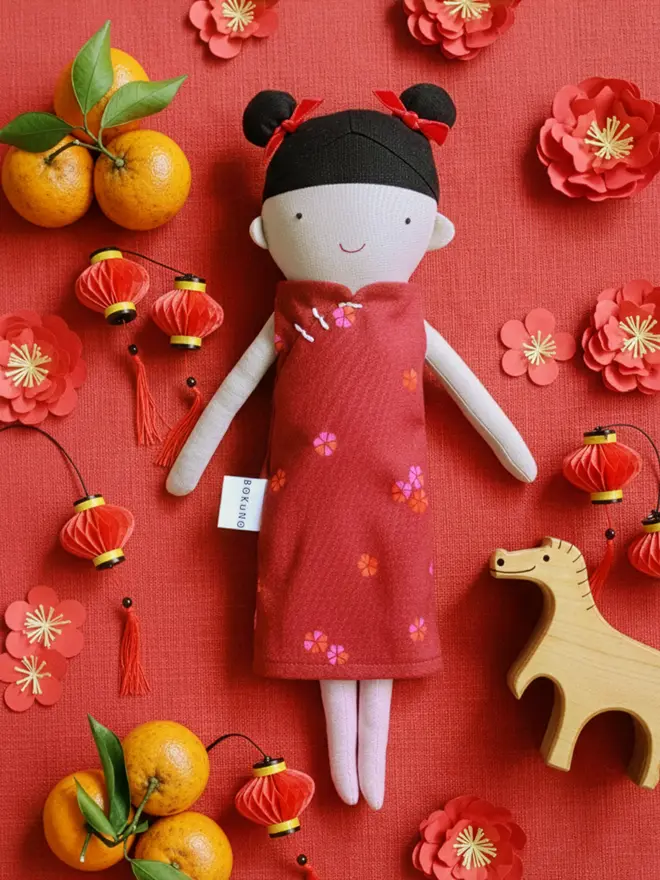 A handmade doll with black hair and red bows, wearing a red dress with floral accents, surrounded by wooden horse, oranges, paper cut flowers and red lanterns for Chinese themed decoration on red background.