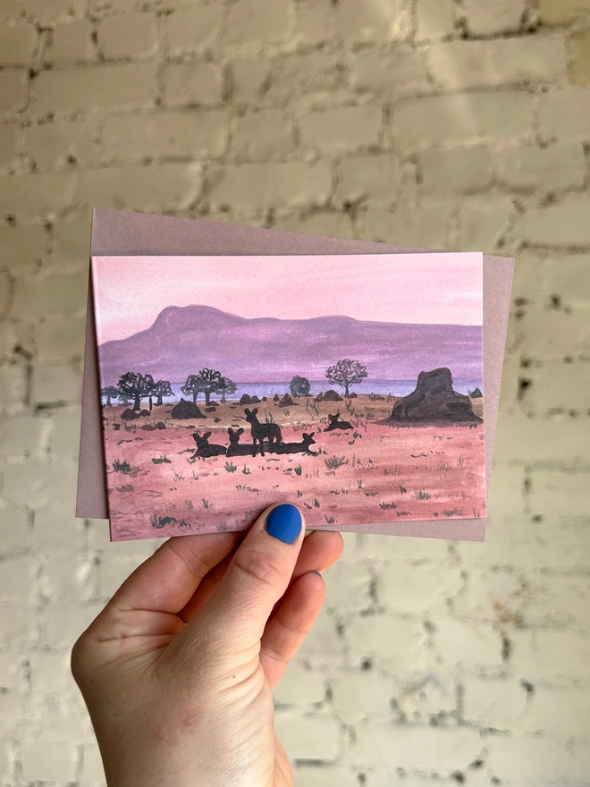 a greetings card featuring an illustration of a dusky coloured landscape and the silhouettes of some african wild dogs with a purple envelope