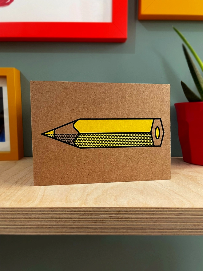 Yellow pencil with black outline and halftone detail, screenpritned on Kraft brown landscape card. Sat on a plywood shelf, with coloured picture frames around and a bit of a plant showing to one side.