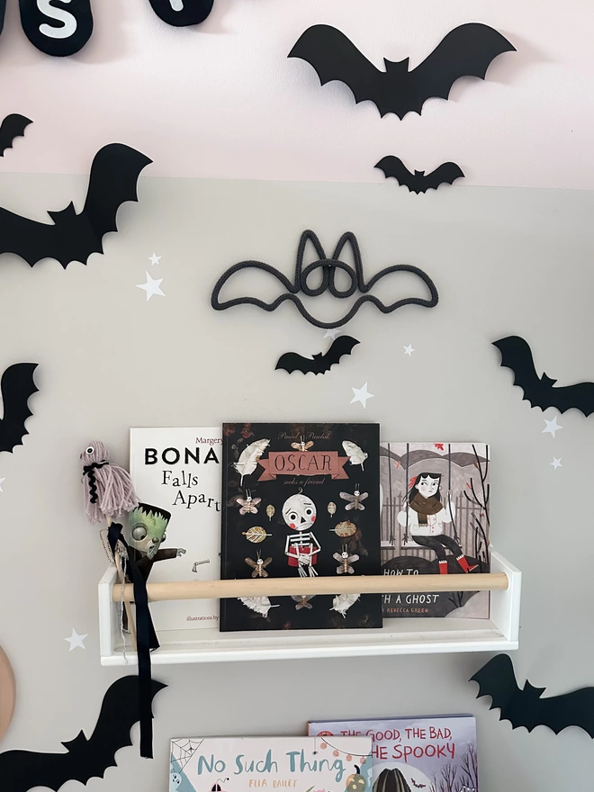 A cute and spooky knitted bat wall hanging, hung up on the wall above a bookshelf decorated for Hallow's Eve.