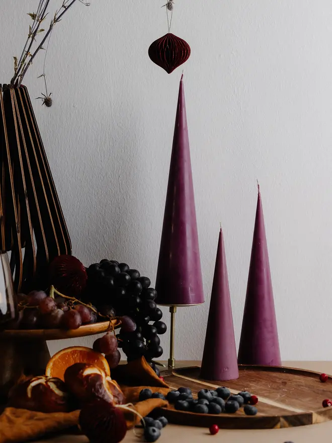 Burgundy Christmas Tree Cone Candles (Set Of 3). Three plum cone candles in tall, medium and small sizes on a wooden tray with blueberries and cranberries, next to grapes, orange slices and a hanging paper ornament. The minimalist tapered candles make a modern, eye-catching addition to seasonal styling.