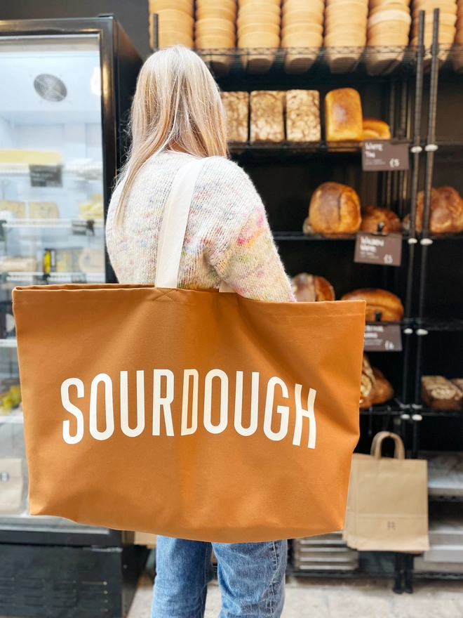 model carrying an oversized tan canvas tote bag with sourdough slogan in a bakery