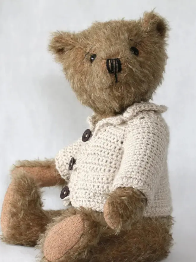 Brown teddy bear sitting upright, wearing a cream knitted cardigan with three buttons, against a plain light background.