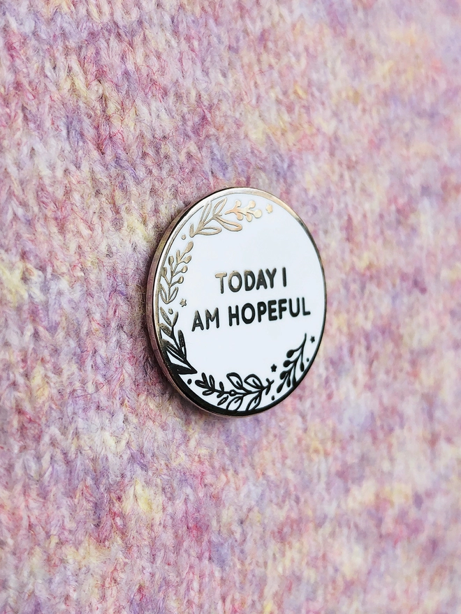 A round white enamel pin with a floral design and the words "Today I Am Hopeful" is pinned to lilac fabric.