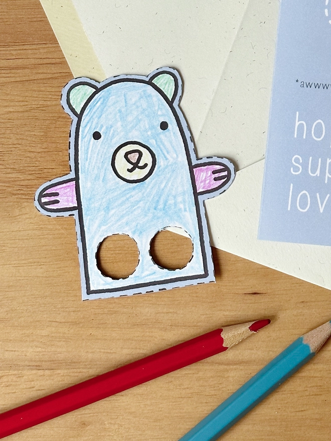 A colour in Christmas card with a polar bear finger puppet design lays on a white envelope on a wooden desk.