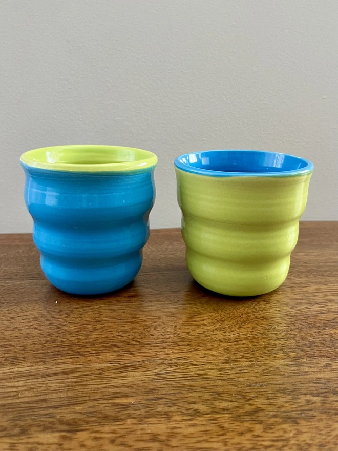 a pair of ceramic tumblers with a wavy sides on a wooden table. the pair is glazed in reverse colours. one is azure blue on the outside and lime green on the inside and the other is the opposite with the lime green on the outside and azure blue on the inside.  