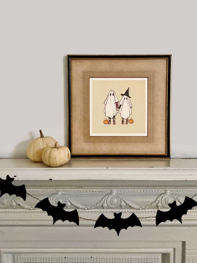 a halloween print of 2 cute ghosts on a mantle piece with a bat garland and 2 pumpkins