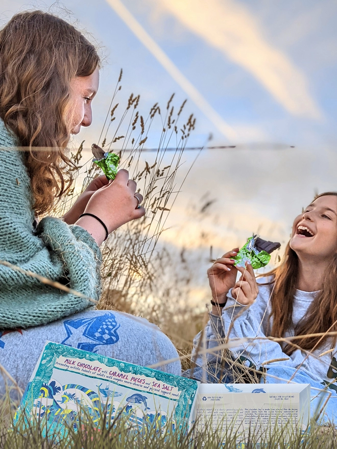 Two happy girls in a field enjoying charity milk chocolate wrapped in green foil & whale packaging  