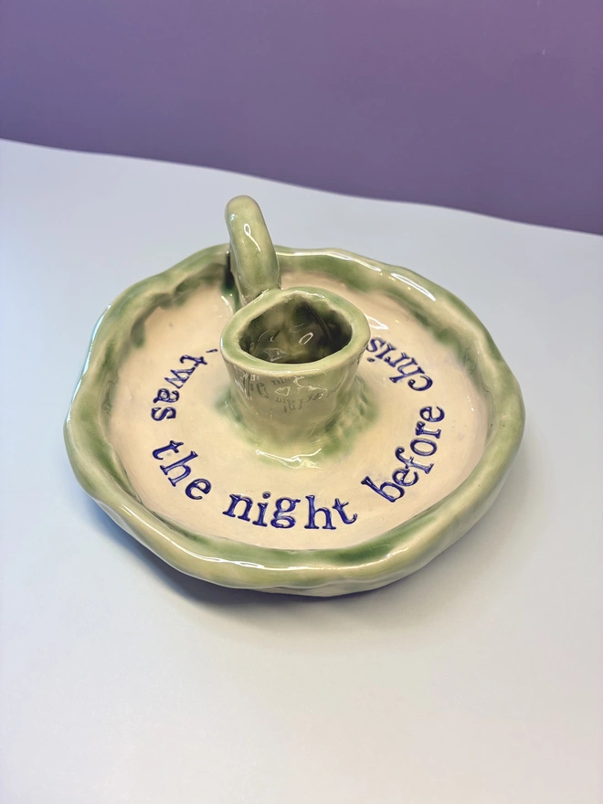 Handmade ceramic Christmas taper candlestick holder with a soft green glazed edge and the words “‘Twas the night before Christmas” in dark blue lettering, arranged around the candle holder opening. Photographed against a pale background.