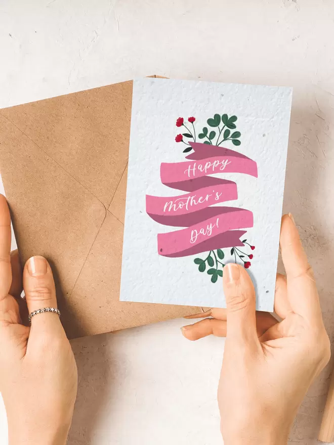 A pair of hands holding a card and brown envelope. The card has a 'Happy Mother's Day' design on it.