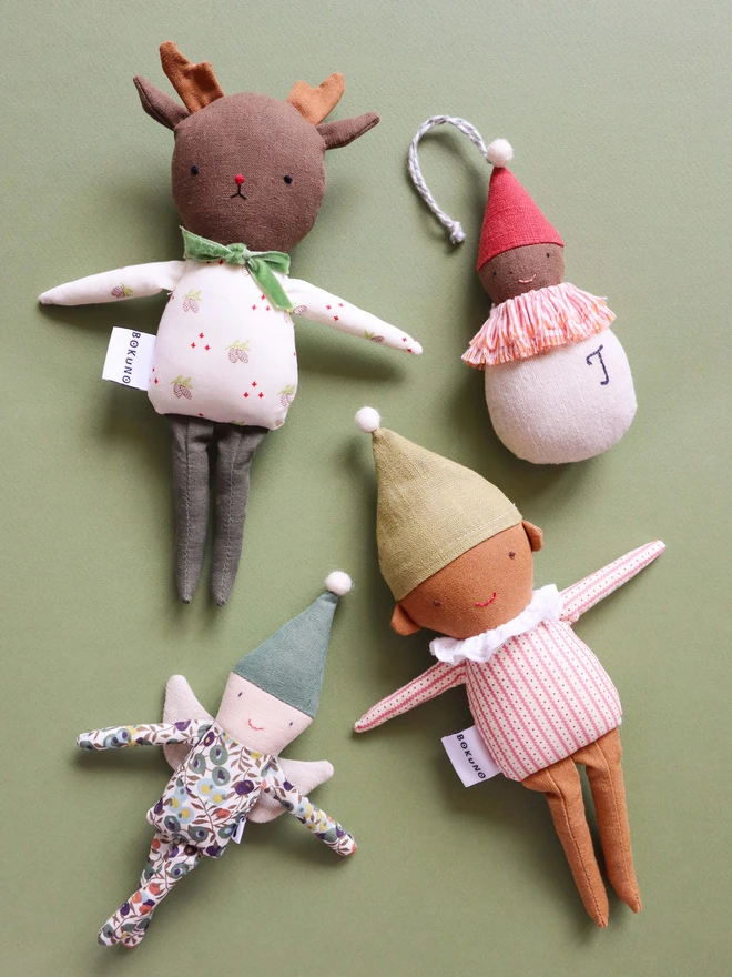 A reindeer doll with soft antlers, embroidered facial features, and a festive pinecone print outfit with either green velvet bow, arrnaged on a green backgroumd with other christmas themed dolls.