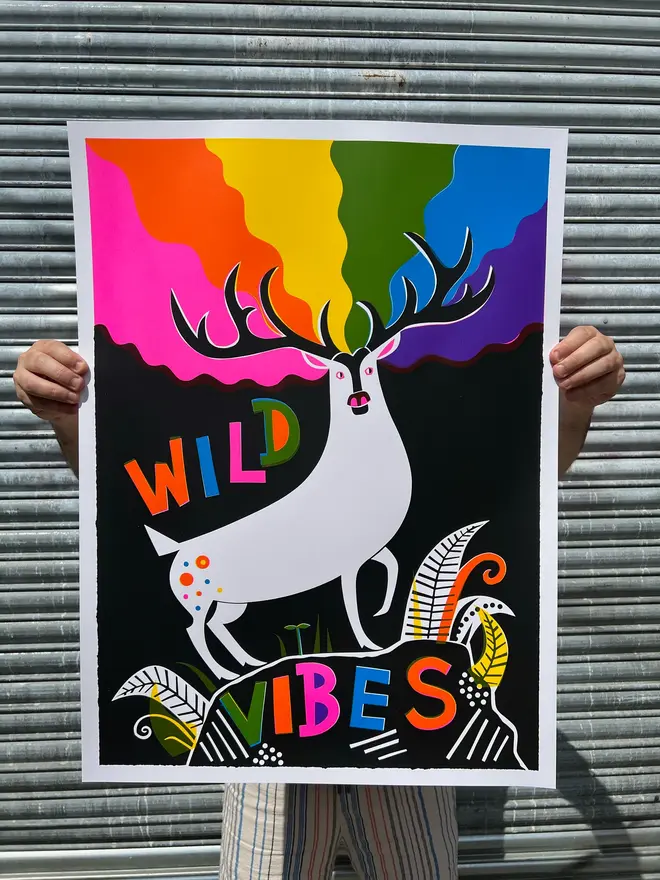 'Wild Vibes' Screenprint, a person holding a colourful screenprint against a metal shutter. The print reads 'wild vibes'. 