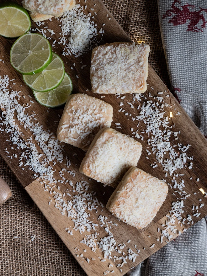 Lime and coconut welshcakes on a wooden chopping board surrounded by desiccated coconut and sliced lime 
