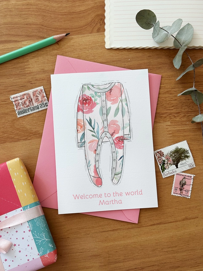 An illustrated new baby greetings card with a pink onesie design lays on a pink envelope on a wooden desk.