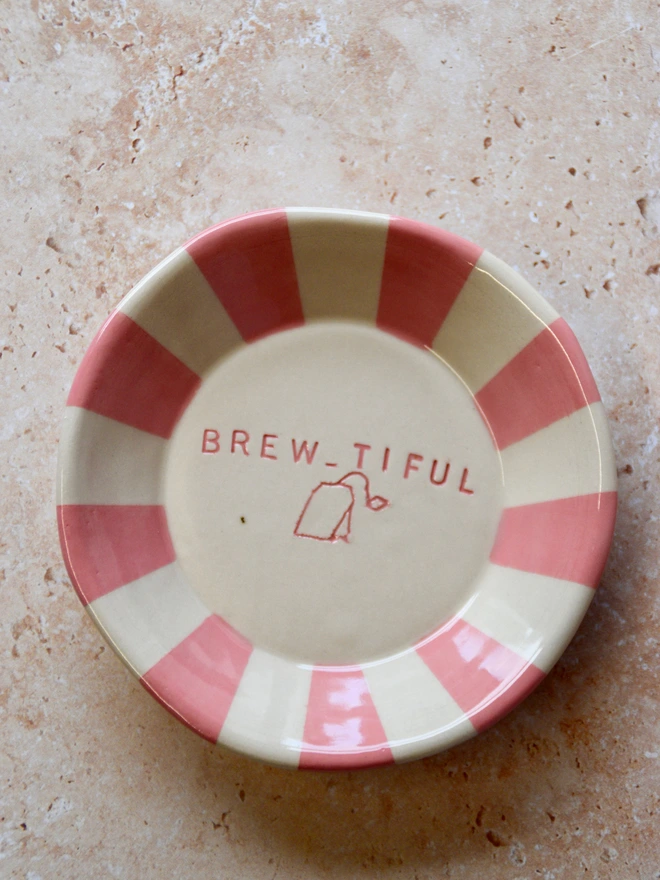 Handmade ceramic tea bag dish with pink striped rim and “Brew-tiful” text and teabag illustration in the centre, styled on a neutral stone background.