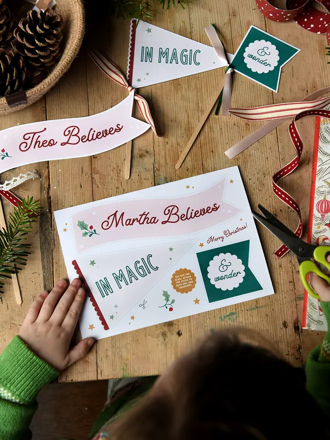 A Christmas card featuring three personalised flags that can be cut out and crafted. The flags read "Martha believes in magic & wonder" and the card lays on a wooden table.