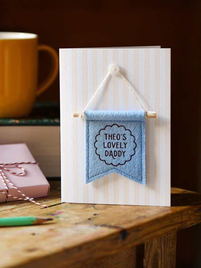 Handmade Father’s Day card with personalised blue felt banner “Theo's Lovely Daddy”, miniature hanging flag keepsake on wooden table. The flag is hung on the card from a tiny mother of pearl button. Beside the card is a book, a mug, a wrapped gift.