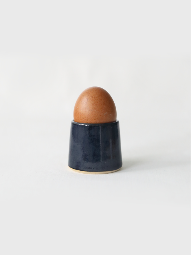A handmade ceramic egg cup in jet black glaze holding a brown egg, photographed against a plain white background