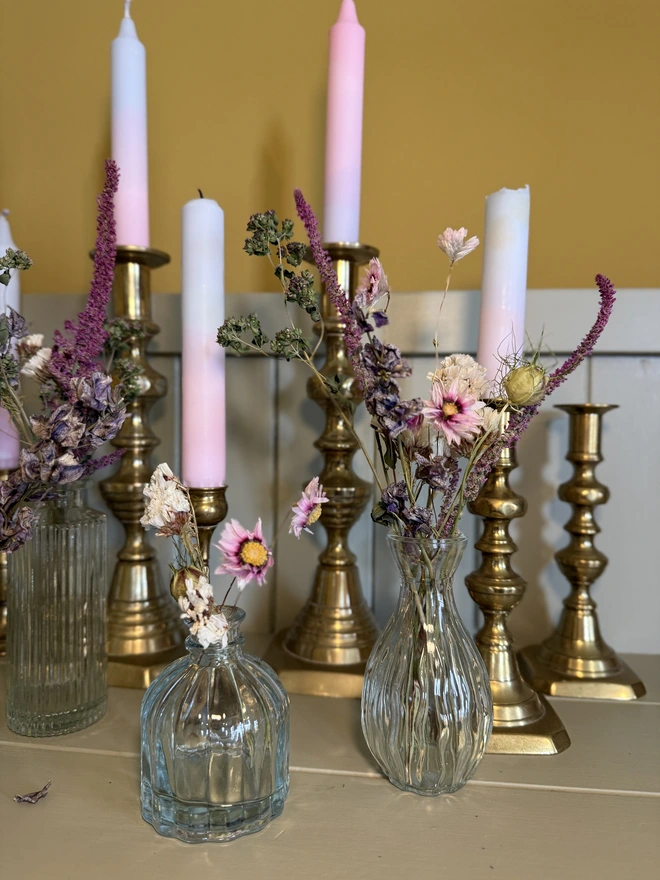 a selection of dried flowers in glass bud vases on a shelf with candles in brass candlesticks