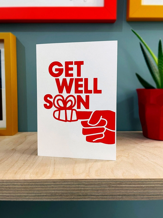  A finger wrapped in a bandage form the two o’s of the word ‘soon’ as part of this Get well soon card. Printed in red ink on a white portrait format card. Stood on a plywood shelf with hints of coloured frames around and a plant to one side.  