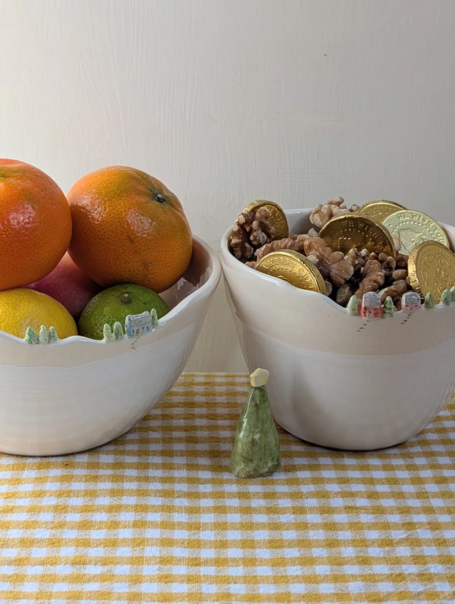 Two Handmade ceramic white bowls with tiny houses and tree details on the rim and white glaze on a yellow gingham table cloth