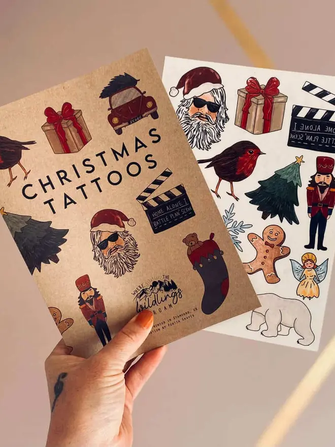 Christmas Temporary Tattoos, a hand holds a brown kraft paper booklet titled "CHRISTMAS TATTOOS" next to a sheet of festive holiday temporary tattoos.