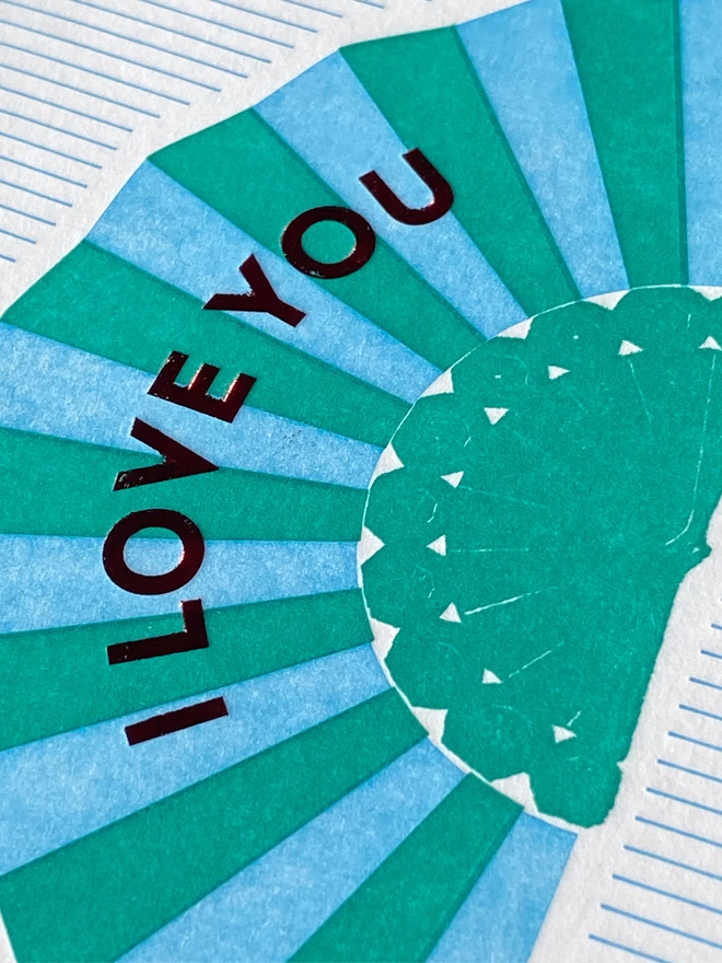 Close up of a White card with blue and green fan with red text reading 'I Love You' on green and white striped card.