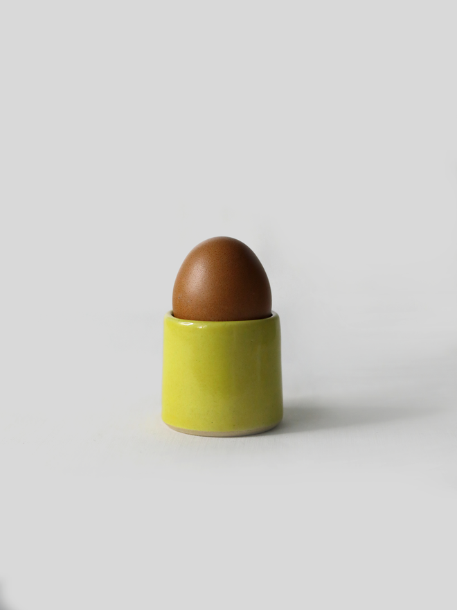 A handmade ceramic egg cup in sun yellow glaze holding a brown egg, photographed against a plain white background