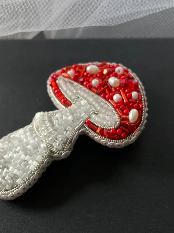 Mushroom Beaded Brooch displayed on a grey box, next to some white fabric. 