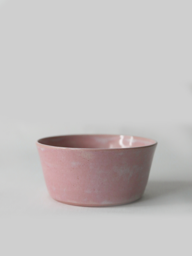 A single hand-thrown ceramic breakfast bowl in blush pink, with a glossy glaze showing subtle variation in tone, a gently flared form and unglazed natural clay base, photographed against a plain light grey background.