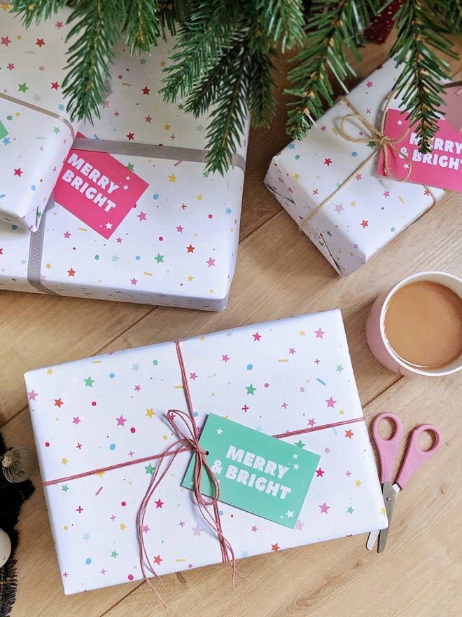 Gifts wrapped in pastel star Christmas wrapping paper are on a wooden floor beneath a Christmas Tree.